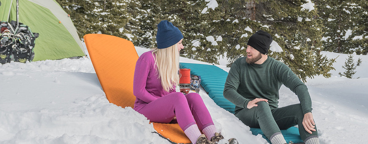 Wide front view of a woman in a magenta merino wool base-layer set and navy beanie sitting in snow with a mug beside a man in a forest green base-layer set and black beanie, with sleeping pads and a tent behind them