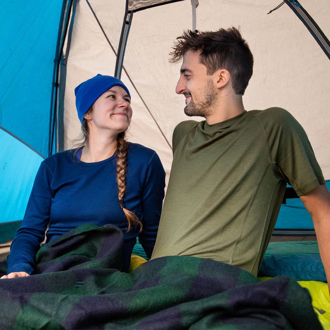 Two people lay together under a wool blanket in a camping tent. The man models the microweight merino wool t shirt 