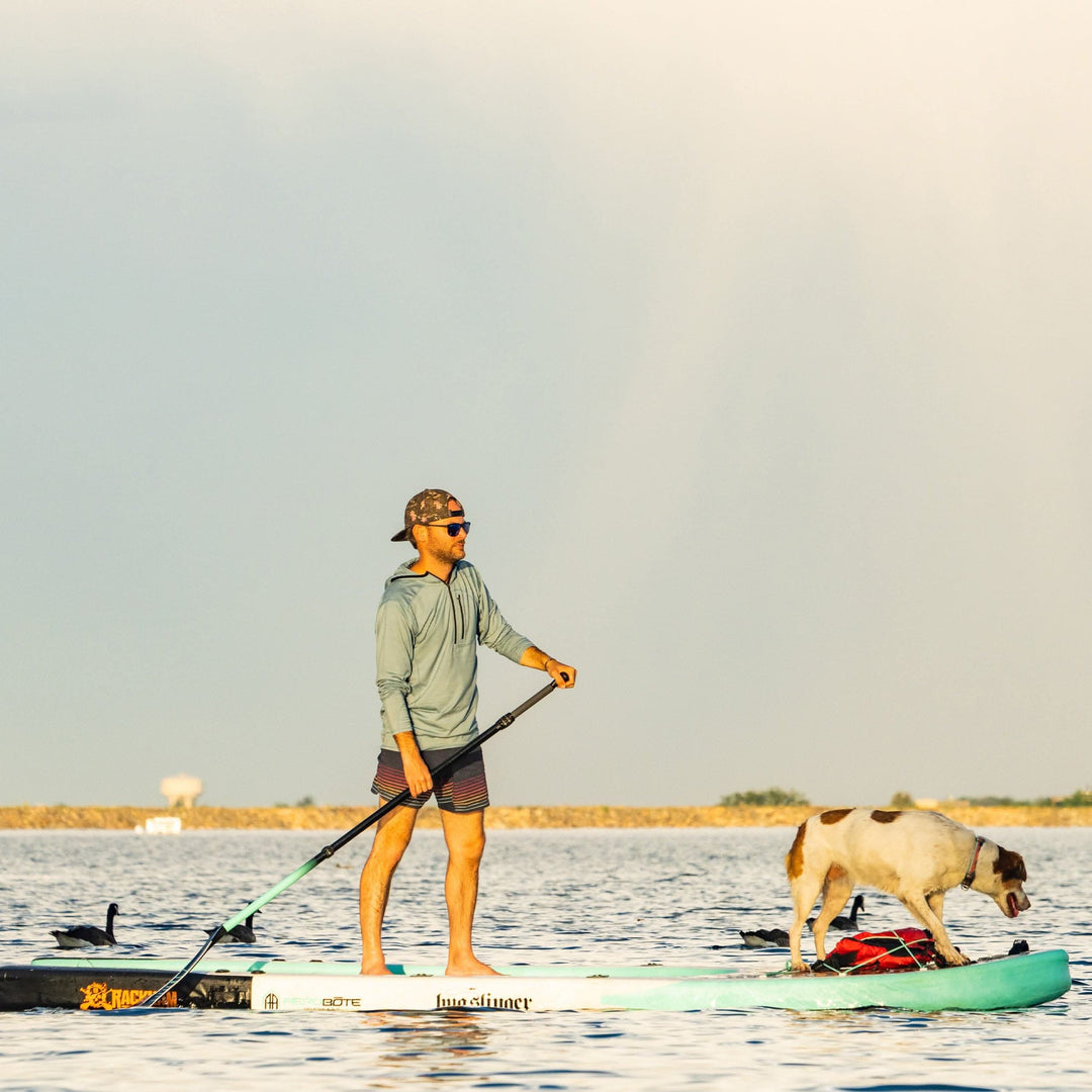 A man paddleboards at sunset with his dog while wearing the super light microweight merino wool quarter zip sun hoodie. 