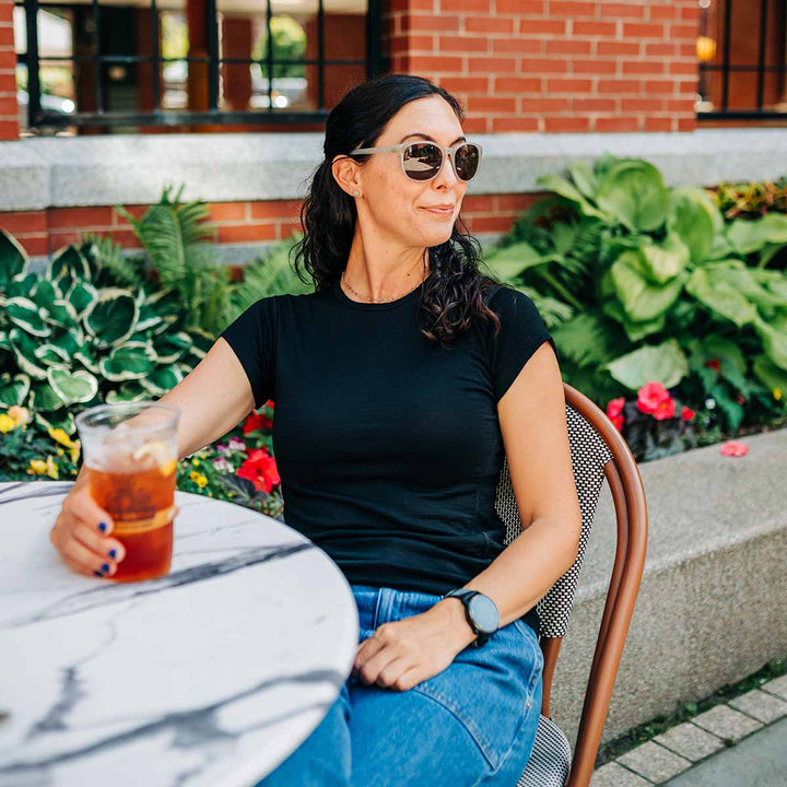 Woman sitting outdoors with a drink, wearing sunglasses and a black merino wool shirt. 