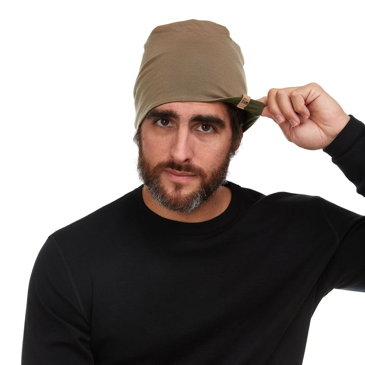 Front three-quarter view of a male model wearing a tan and olive drab reversible merino wool blend beanie with the cuff turned up 