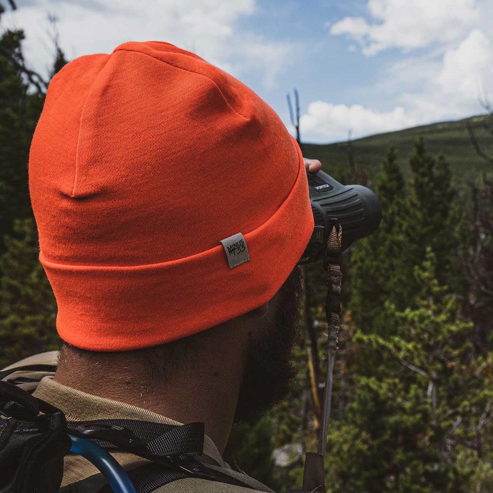 A man models the lightweight, double-layered, hundred percent, super soft merino wool beanie perfect for fall, winter, and spring. This hat is perfect for a hike up the mountain or a walk around the block, front three-quarter view outdoors #color_blaze-orange