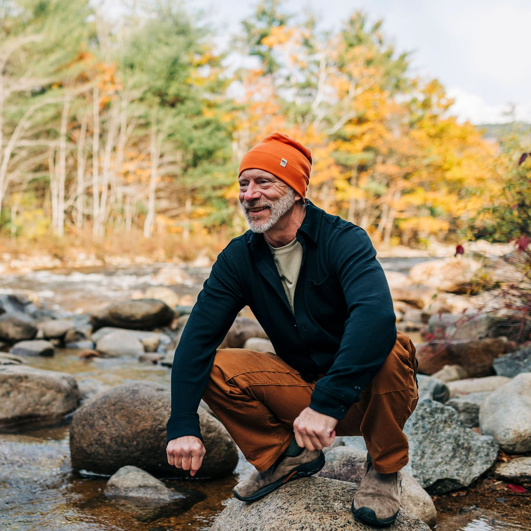 A man squats on a rock while wearing the lightweight, double-layered, hundred percent, super soft merino wool beanie perfect for fall, winter, and spring. This hat is perfect for a hike up the mountain or a walk around the block!
