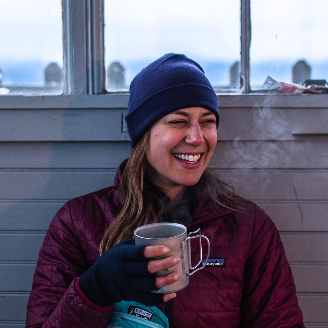 Person wearing a blue knit beanie and maroon jacket sits indoors holding a steaming mug.