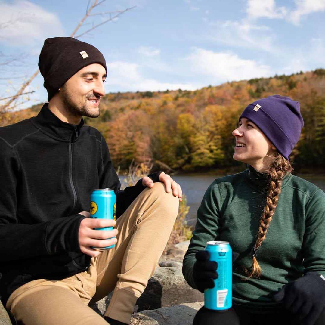 Two people sit on a rock engrossed in conversation in front of a mountain-lined lake. The man models the lightweight beanie, heavyweight full zip sweater, and midweight merino wool base layer bottoms. The woman models the lightweight beanie, midweight quarter zip mock neck top, and merino wool glove liners. 