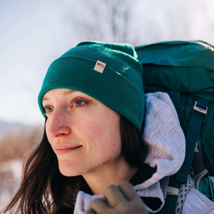 A woman models a lightweight, double-layered, hundred percent, super soft merino wool beanie with our heavyweight brushed wool hoodie on a winter trail. This hat is perfect for a hike up the mountain or a walk around the block!
