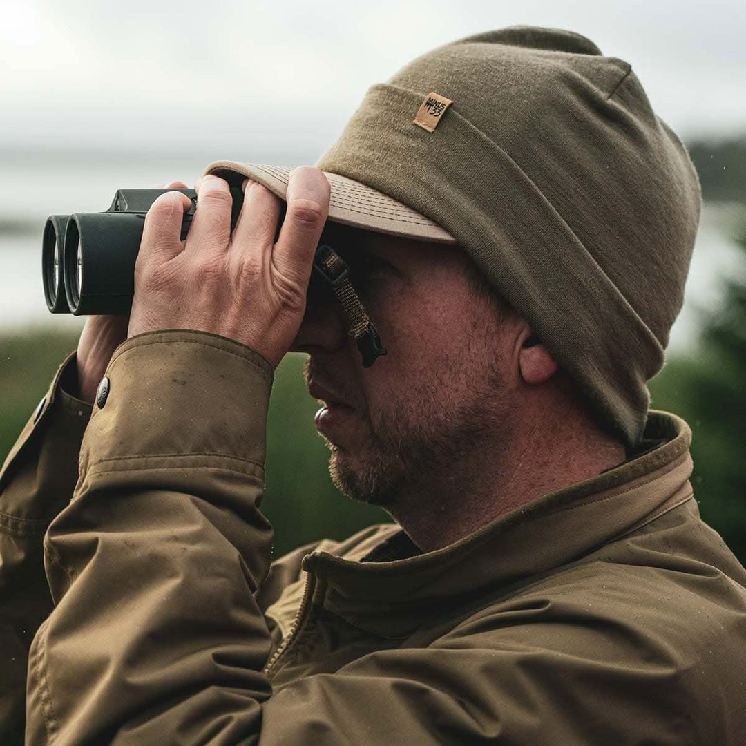 A man models the lightweight, double-layered, hundred percent, super soft merino wool beanie over a baseball cap while looking through binoculars. This hat is perfect for a hike up the mountain or a walk around the block!
