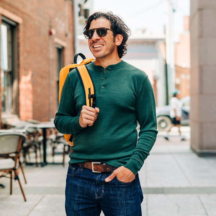 Man wearing a green merino wool sweater and sunglasses, holding a yellow backpack in an urban setting. 