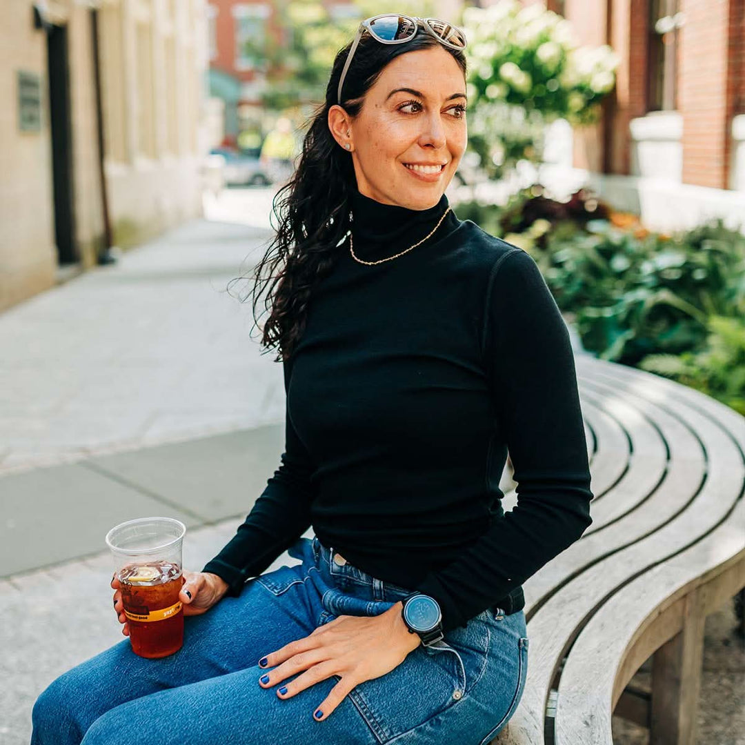 Woman sitting on a bench holding a drink, wearing a black merino wool turtleneck sweater and blue jeans, with a blurred outdoor background. 