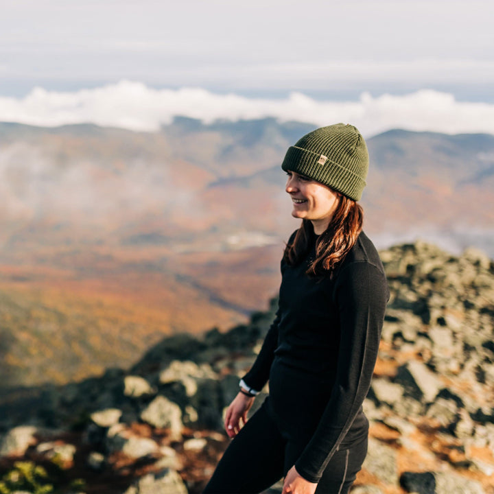 A woman on a mountaintop models the midweight hundred percent merino wool beanie 