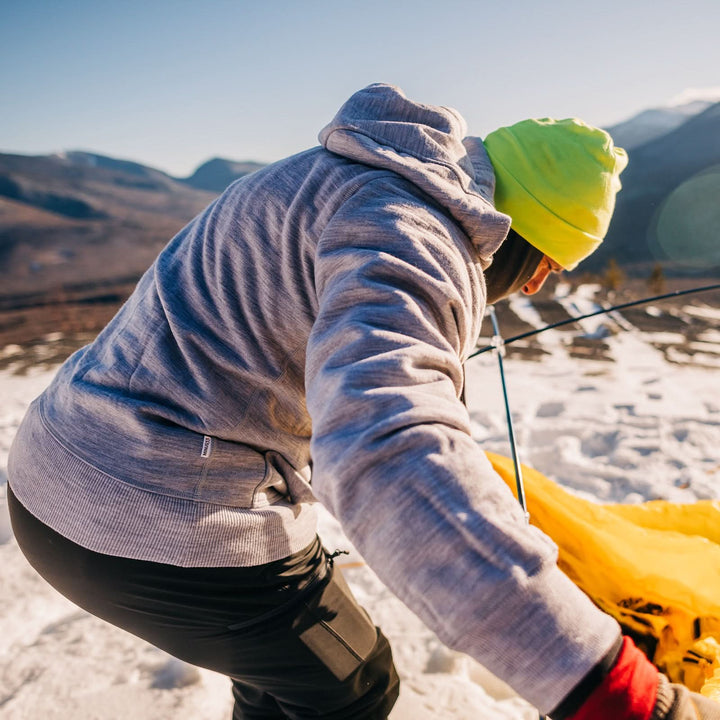 A man sets up his tent on a snowy mountain. The man is modeling the heavyweight brushed wool Kodiak Fleece Hoodie, crafted with thumbholes and a drawstring to bundle you up for wicked cold days! 