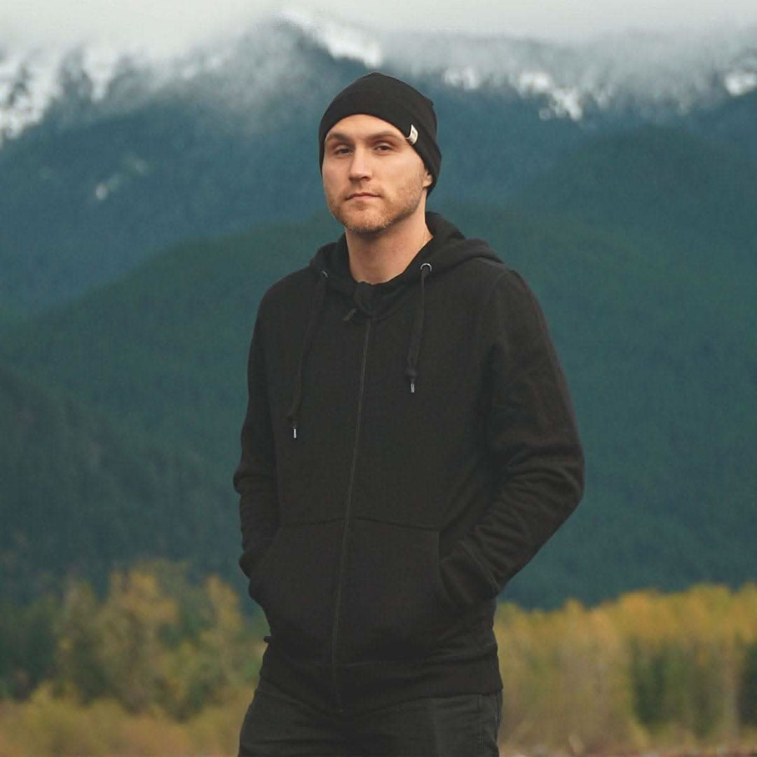 A man stands in front of mountains wearing the heavyweight brushed wool Kodiak Fleece Hoodie, crafted with thumbholes and a drawstring to bundle you up for wicked cold days! 