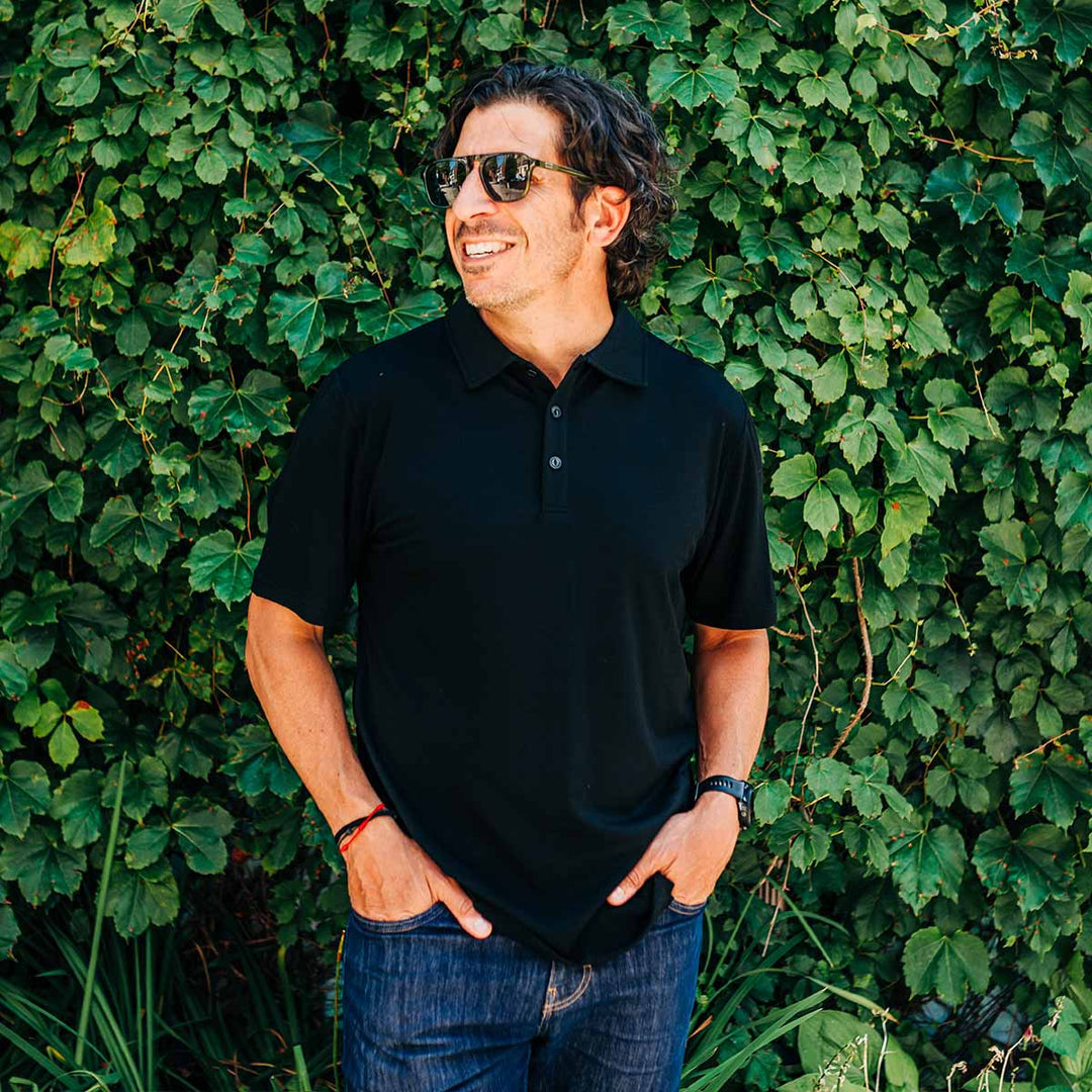 Man wearing a black polo shirt and sunglasses standing against a green leafy background wearing a merino wool polo 