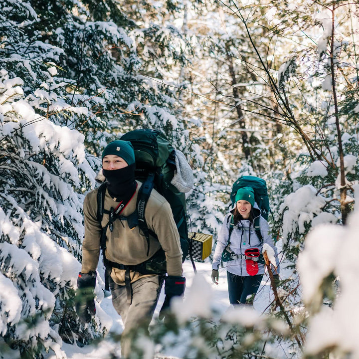 A man models a super soft, long sleeved merino wool thermal base layer crew. This top makes for a perfect first layer on a winter hike, or a nice casual wear on the chilly days of autumn and spring, man in a tan crew top leading another backpacker through a snowy forest 