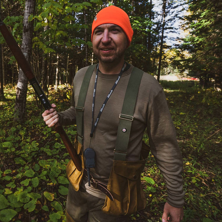 A man models a super soft, long sleeved merino wool thermal base layer crew. This top makes for a perfect first layer on a winter hike, or a nice casual wear on the chilly days of autumn and spring, man in a tan crew top holding a rifle on a wooded trail 