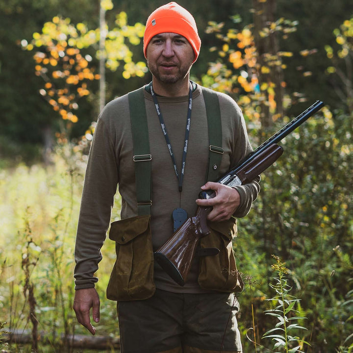 A man models a super soft, long sleeved merino wool thermal base layer crew. This top makes for a perfect first layer on a winter hike, or a nice casual wear on the chilly days of autumn and spring, front view of a man in a tan crew top holding a shotgun at a forest edge 