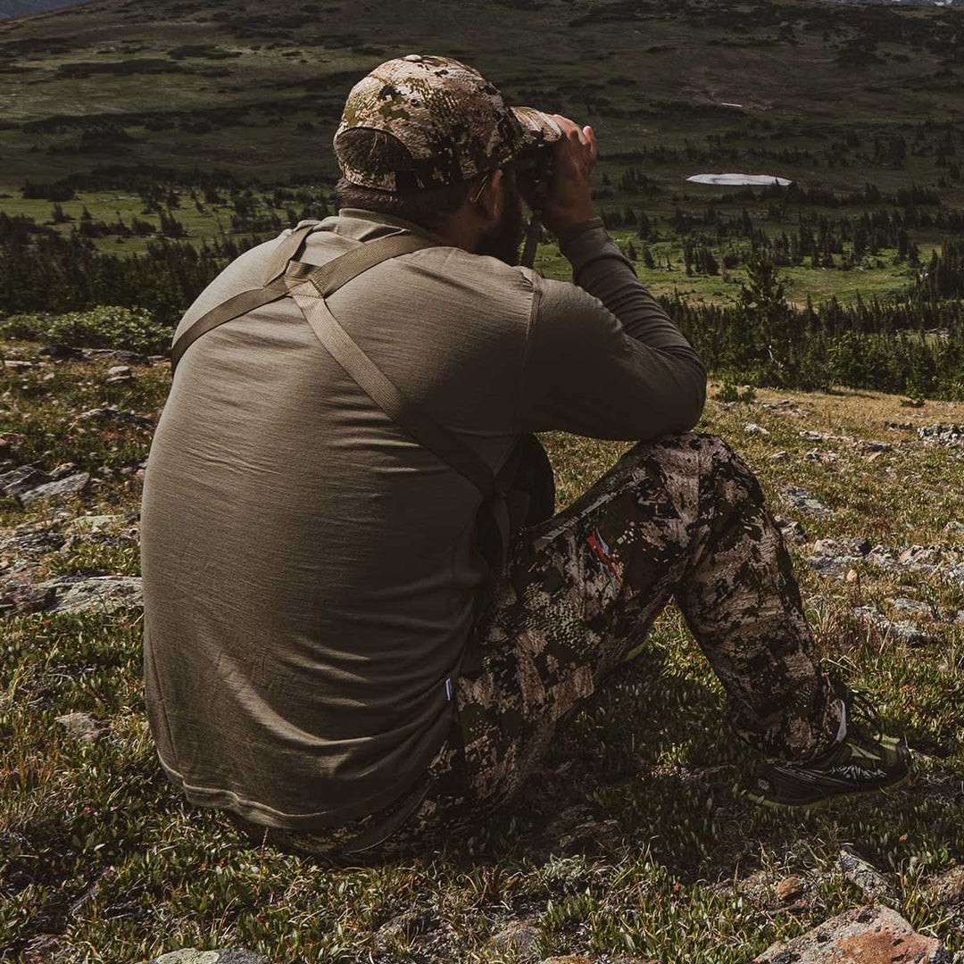 A man sits high on a hill and gazes through binoculars while modeling the lightweight long sleeved merino wool base layer crew neck top 