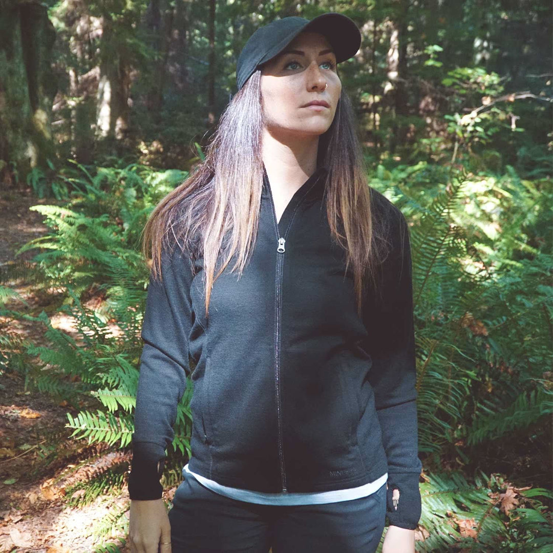 A woman stands in front of tall ferns surrounded by autumn leaves on the ground. Sunlight peeks through the trees. The woman is wearing a hundred percent merino wool heavyweight full zip sweater. This outer layer features a two and a half inch mock neck collar, two zippered pockets, and thumbholes. 