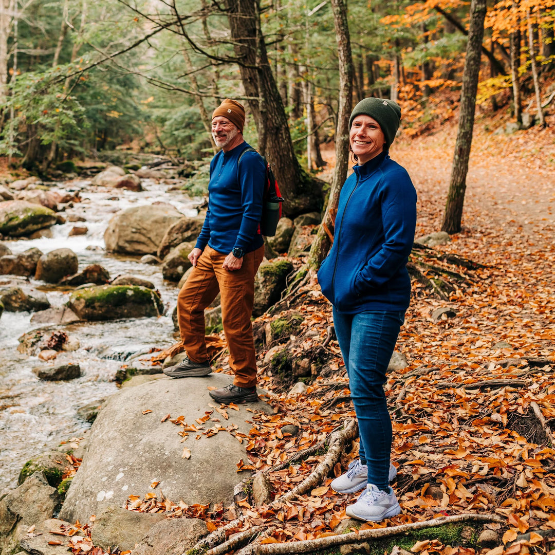 Two people stand on a trail covered in autumn leaves at the edge of a stony, flowing brook. The woman wears a merino wool hat and a hundred percent merino wool heavyweight full zip sweater. This outer layer features a two and a half inch mock neck collar, two zippered pockets, and thumbholes. 