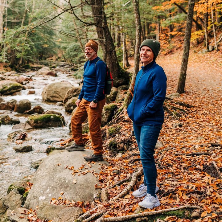 Two people stand on a trail covered in autumn leaves at the edge of a stony, flowing brook. The woman wears a merino wool hat and a hundred percent merino wool heavyweight full zip sweater. This outer layer features a two and a half inch mock neck collar, two zippered pockets, and thumbholes. 