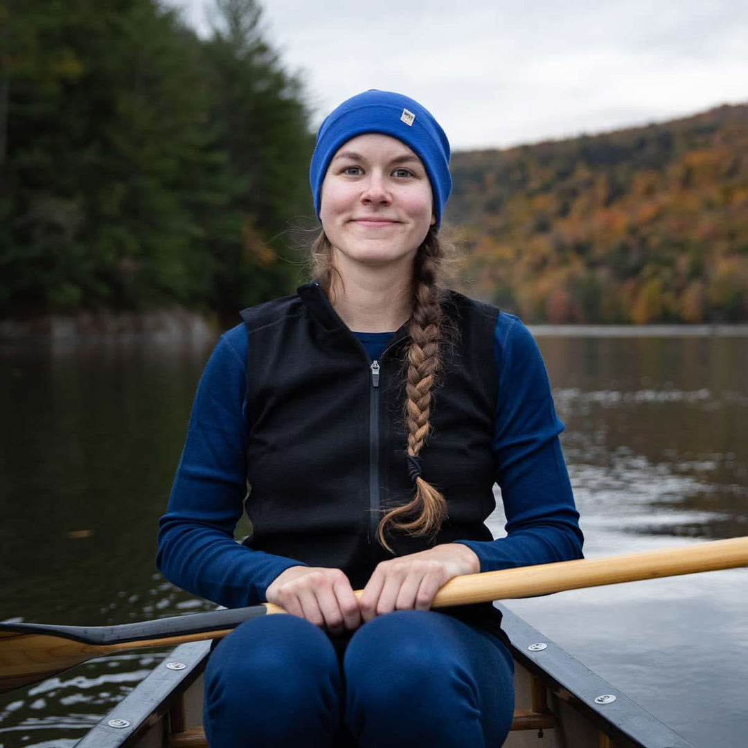 A woman sits in a canoe on open water in front of mountains and trees. The woman is wearing a lightweight hat, midweight base layer top and bottoms, and a hundred percent merino wool heavyweight full zip vest. This outer layer features a two and a half inch mock neck collar and two zippered pockets. 