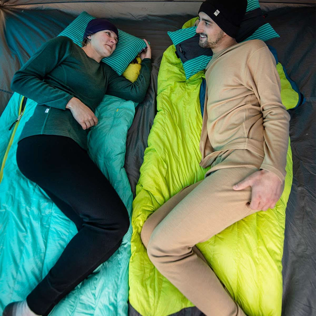 Two people lay on top of sleeping bags in a tent. Both are wearing lightweight hats and midweight base layer tops and bottoms. 