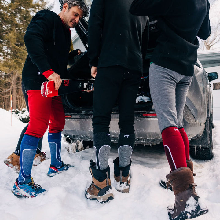 Three people stand in front of a car unloading their skis. The three people each wear a pair of one hundred percent merino wool midweight base layer bottoms, as well as ski and snowboarding socks. These bottoms feature a gusset between the legs for improved range of motion, and cuffs around the ankles for comfort and style. 