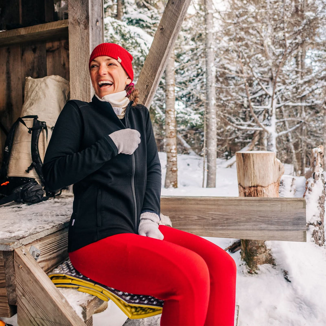 A woman sits on the front step of the camp lodge to adjust her heavyweight full zip sweater. The woman is modeling a pair of one hundred percent merino wool midweight women&