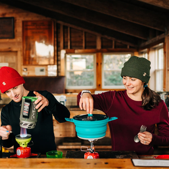 Two people smile while setting up their camping cookware. Both people wear Minus33 merino wool clothing. The woman is wearing the Ossipee midweight one hundred percent merino wool women&