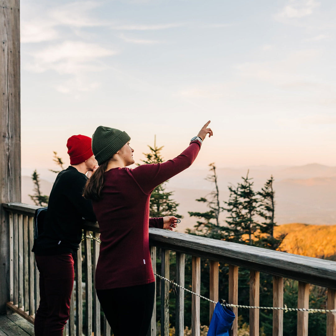 Two people stand on a porch deck watching the sun set below the mountains. Both people wear Minus33 merino wool clothing. The woman is wearing the Ossipee midweight one hundred percent merino wool women&