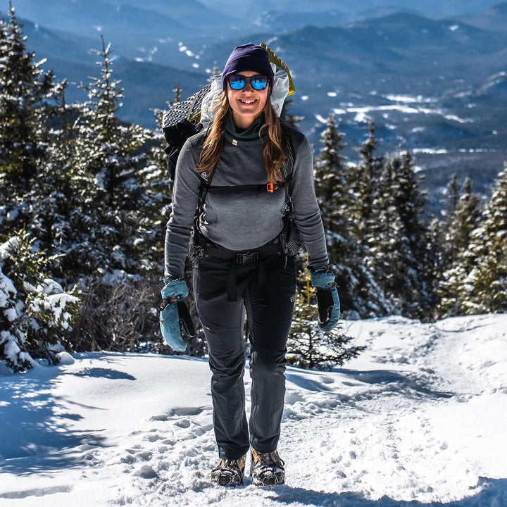 A woman models a soft, 100% merino wool, midweight base layer crew top. She wears a backpack, hat, gloves and sunglasses while hiking a snowy summit. The women&