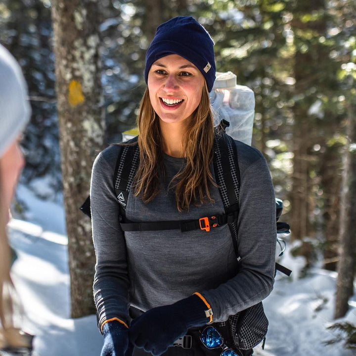 A woman models a soft, 100% merino wool, midweight base layer crew top. She wears a backpack, hat, and gloves while hiking through the snowy forest. The women&