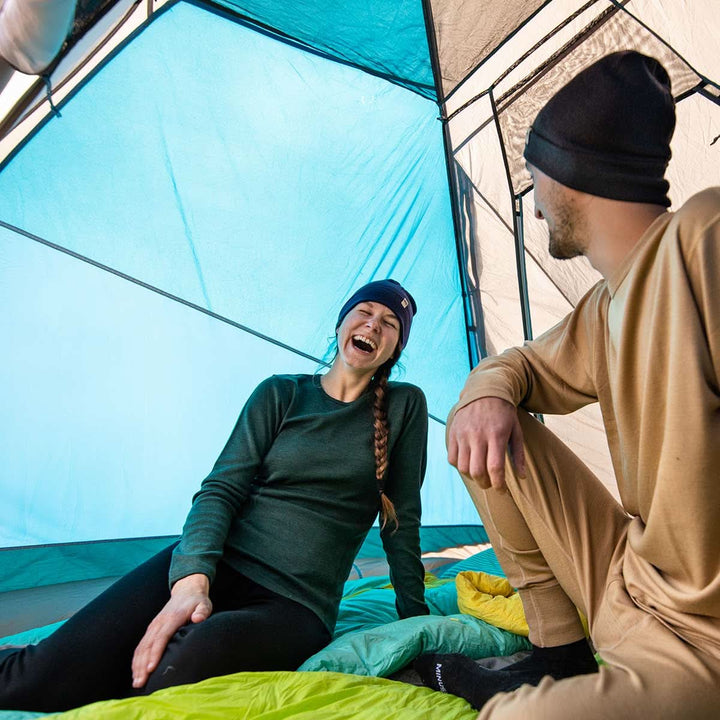 Two people sit in a tent laughing in their super soft one hundred percent merino wool base layers. The woman is wearing the women&