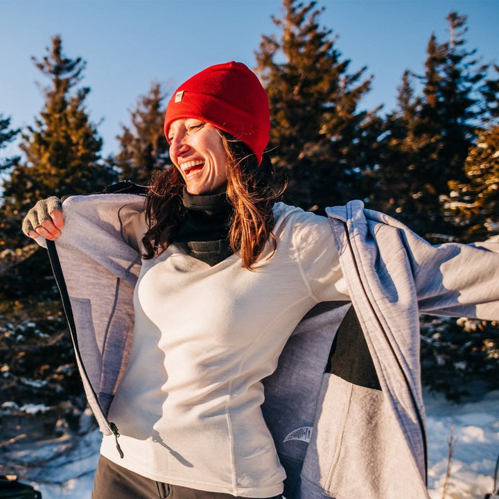 A woman smiles while adjusting her heavyweight merino wool hoodie. The woman is wearing a soft, 100% merino wool, midweight base layer crew top. The women&