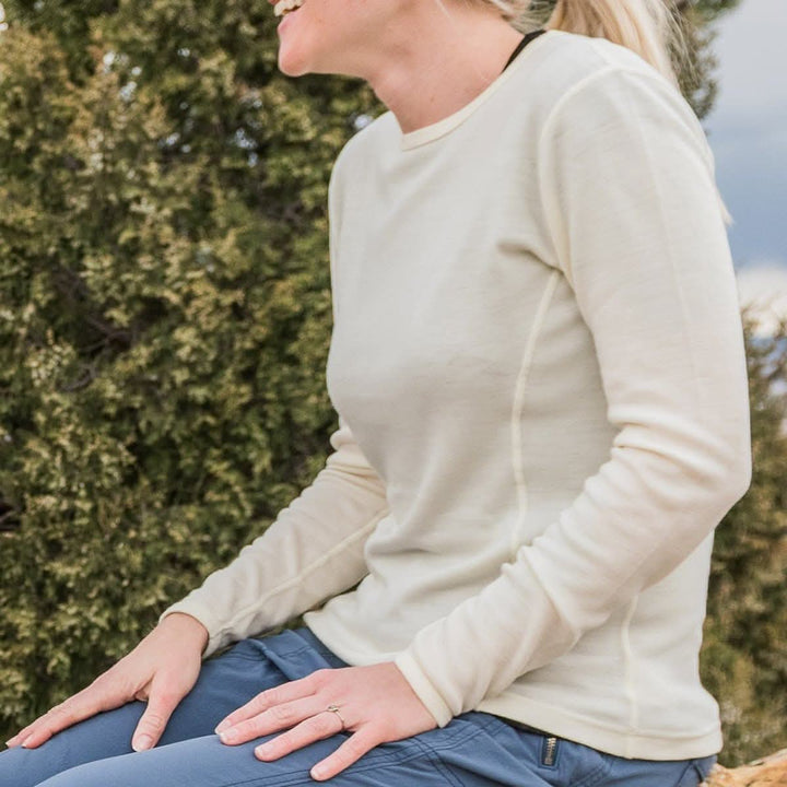 Close up on a woman sitting, wearing a soft, 100% merino wool, midweight base layer crew top. The women&