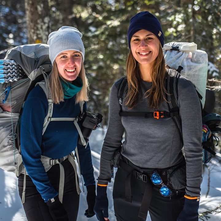 Two women model soft, 100% merino wool, midweight base layer crew tops. They wear backpacks, hats, gloves and a neck gaiter while hiking through the snowy forest. The women&