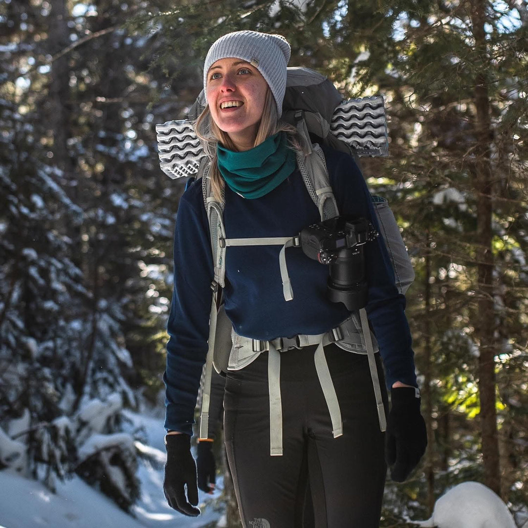 A woman models a soft, 100% merino wool, midweight base layer crew top. She wears a backpack, hat, gloves, and a neck gaiter while hiking through the snowy forest. The women&