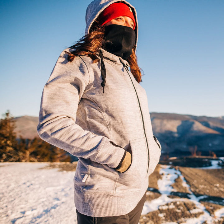 A woman stands in the snow looking out  toward the sun. The woman wears the heavyweight merino wool brushed fleece hoodie, a neck gaiter, and a lightweight beanie. The hoodie features a large hood with drawstrings, thumbholes, two pockets, and an interior stash pocket inside. 