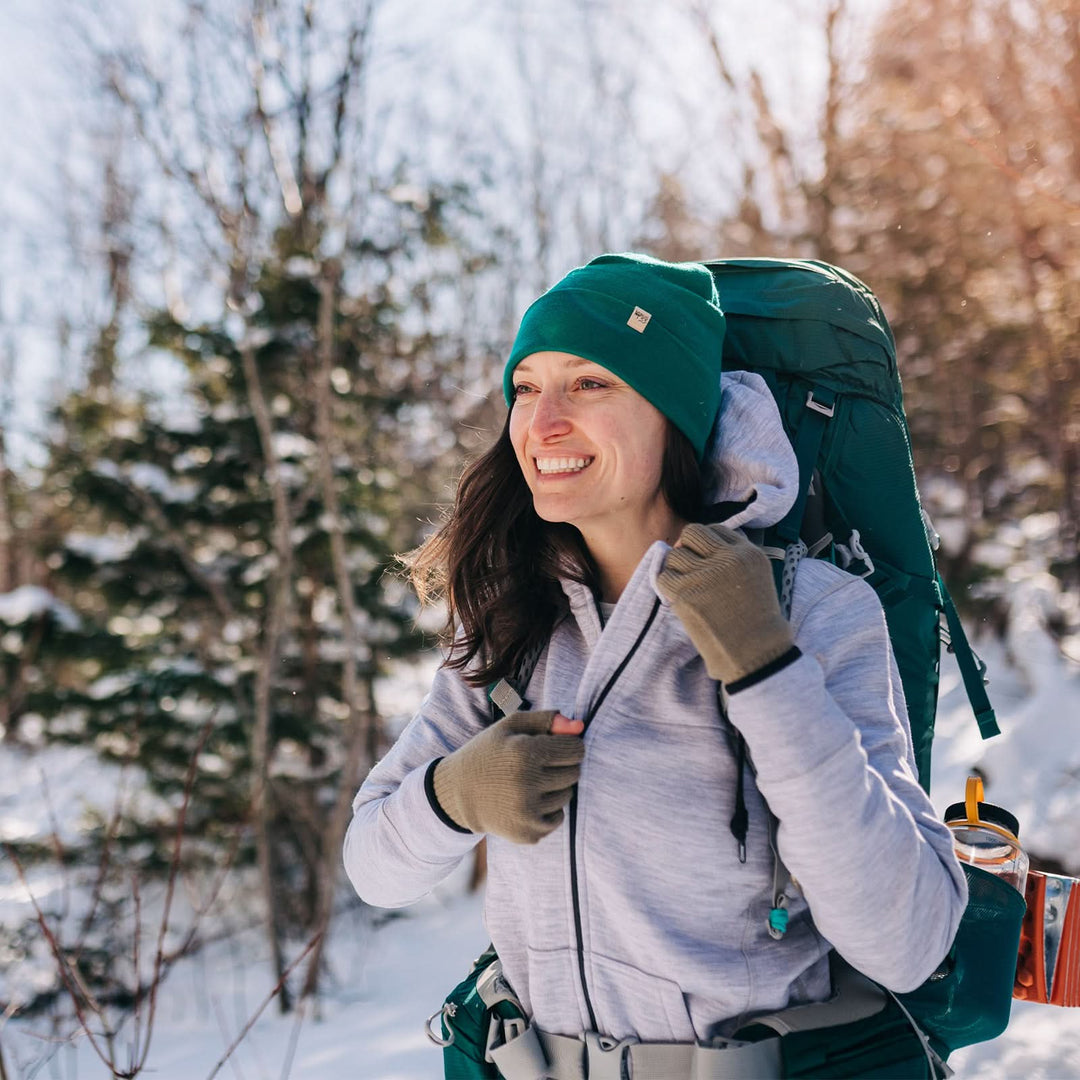 A woman hikes through the snowy forest wearing a backpack over the heavyweight merino wool brushed fleece hoodie. The hoodie features a large hood with drawstrings, thumbholes, two pockets, and an interior stash pocket inside. 