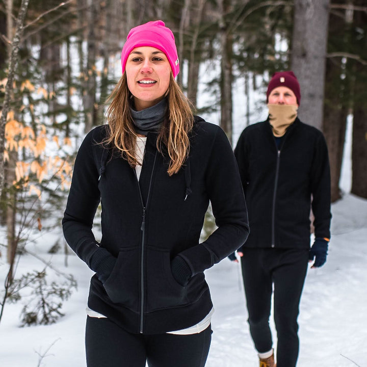 Two people walk through the snowy forest wearing their merino wool gear to keep warm. A woman models the heavyweight merino wool brushed fleece hoodie with a beanie and neck gaiter. The hoodie features a large hood with drawstrings, thumbholes, two pockets, and an interior stash pocket inside. 