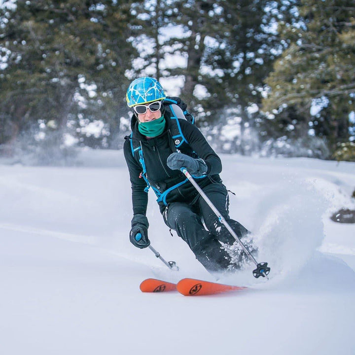 A woman models the heavyweight merino wool brushed fleece hoodie while skiing the slopes. The hoodie features a large hood with drawstrings, thumbholes, two pockets, and an interior stash pocket inside. 