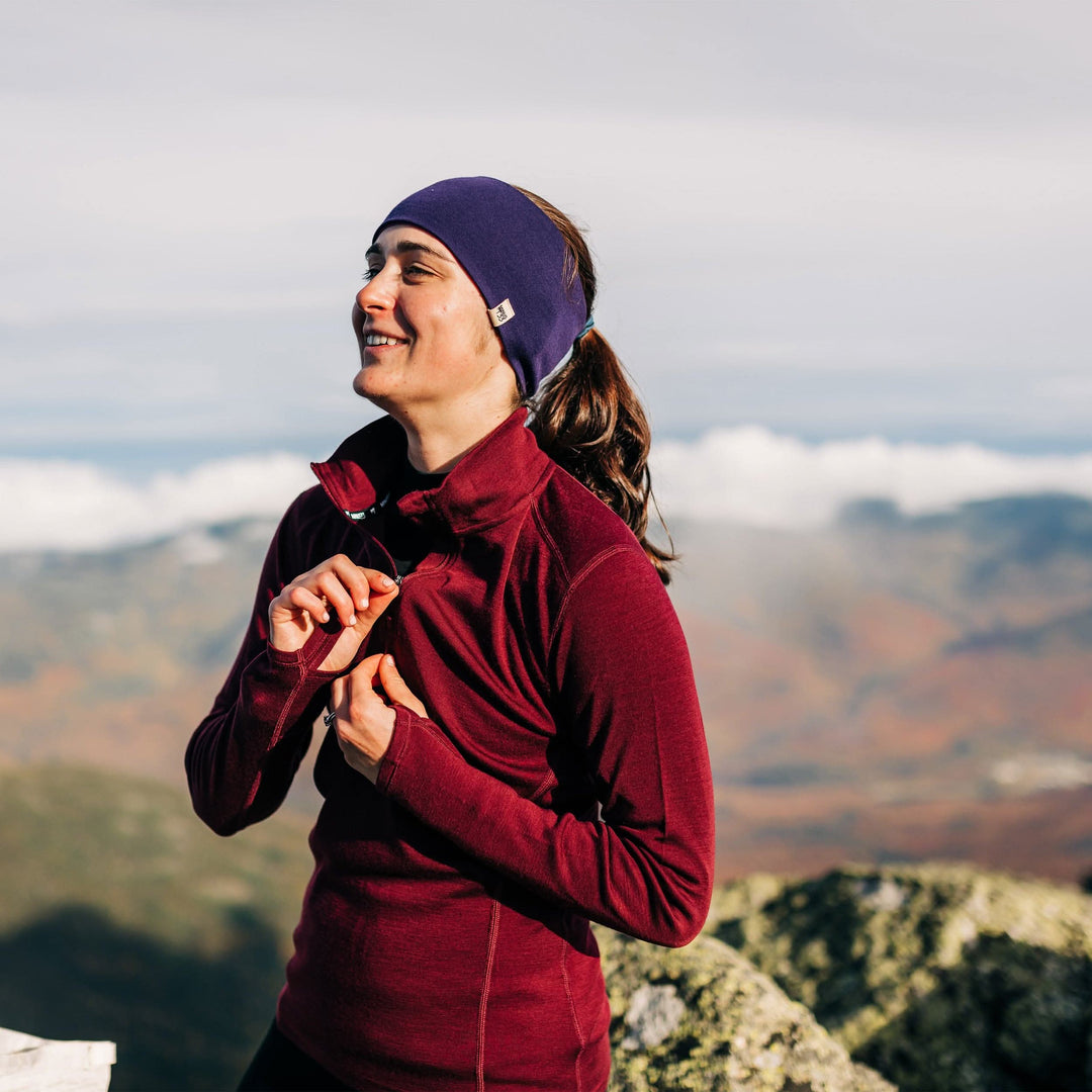 A woman stands on a mountain summit, toggling the zipper on her soft midweight merino wool quarter zip sweater. The sweater features a 1/4 zip neckline and thumbholes at the cuffs 