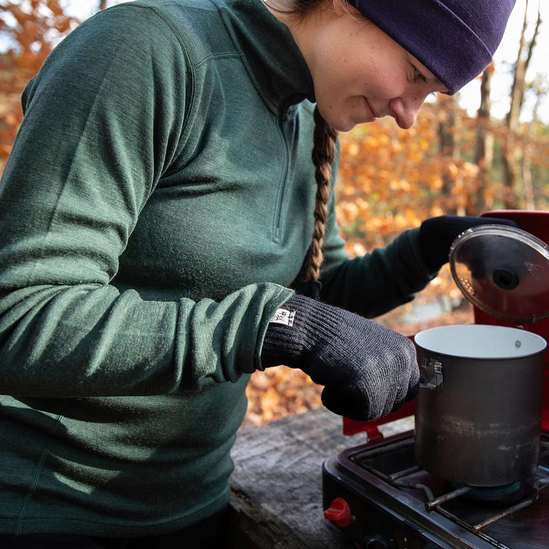 A woman wears her soft merino wool quarter zip sweater while cooking outdoors. The sweater features a 1/4 zip neckline and thumbholes at the cuffs 