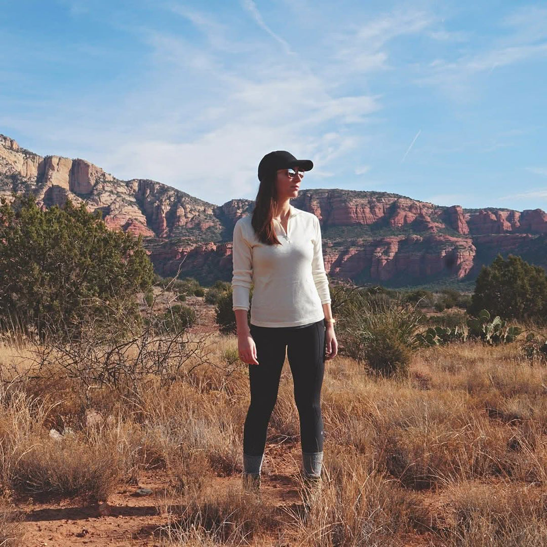 A woman stands in a dry desert field in front of rocky mountains and a blue sky. The model is wearing a one hundred percent merino wool midweight quarter zip sweater. 