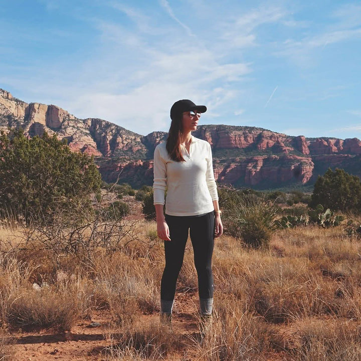 A woman stands in a dry desert field in front of rocky mountains and a blue sky. The model is wearing a one hundred percent merino wool midweight quarter zip sweater. 