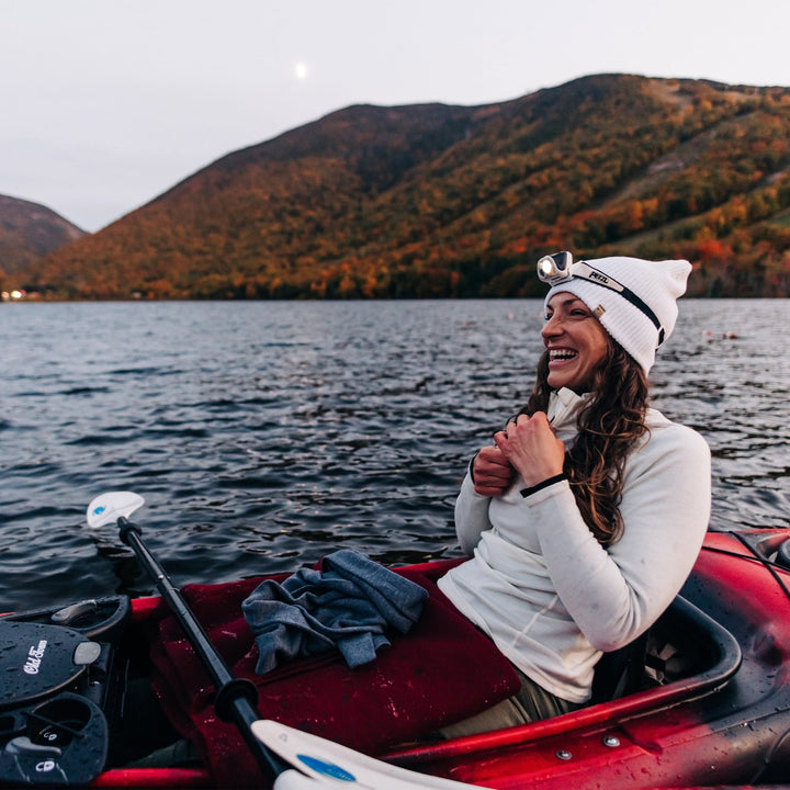 A woman kayaks in the middle of a lake with a backdrop of autumn mountains. The model is wearing a one hundred percent merino wool quarter zip sweater: exceptionally soft, warm, and breathable 