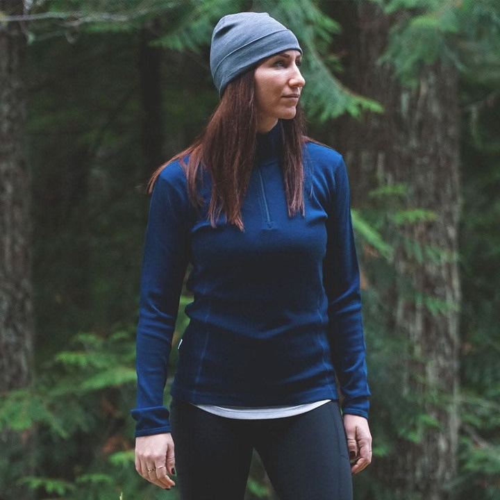 A woman stands in the woods. The model is wearing a one hundred percent merino wool quarter zip sweater with thumbholes 