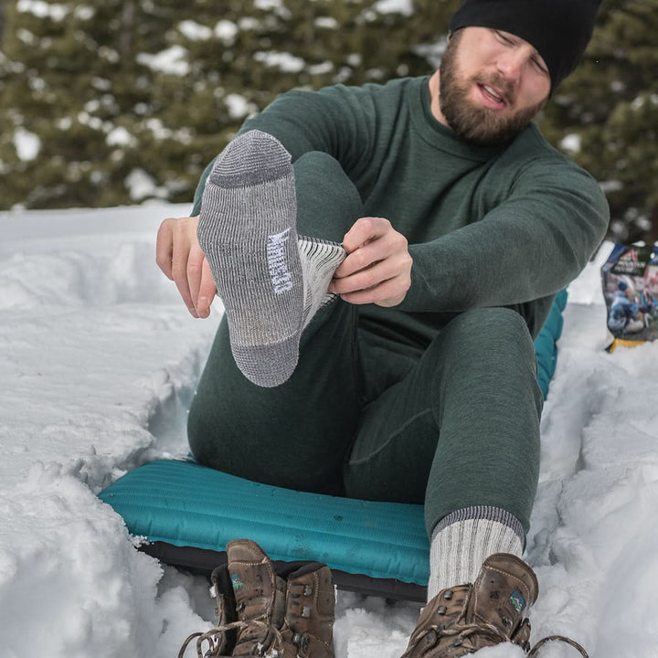 Midweight - Day Hiker Crew Socks - Minus33 Merino Wool Clothing, man pulling on a gray sock while seated in snow