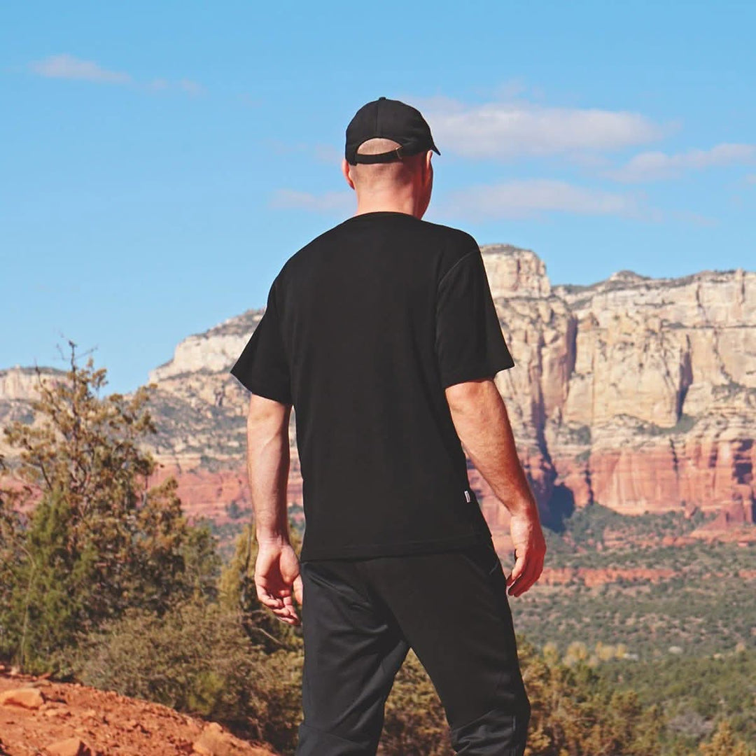 Back view of a man wearing a black Sentinel midweight merino wool short-sleeve crew shirt with black pants and a baseball cap, standing on a red rock trail and looking toward layered canyon cliffs