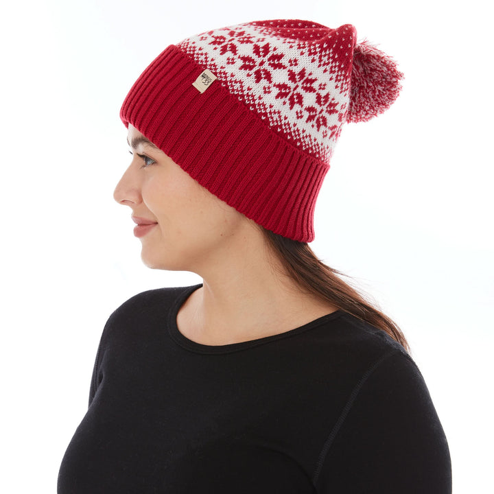 Close-up left side profile view of a woman wearing a true red midweight merino wool cuffed beanie with a natural cream snowflake knit pattern, logo patch, and red-and-cream pom-pom 